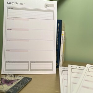 May include: A white daily planner with grey headings and lines for writing. The headings include "Quick Wins", "Appointments", "Meetings", "Tasks", "Evening", "Non-Priority Tasks", and "Referred". The planner has a space for the date at the top.