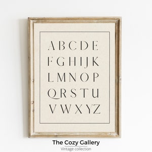 May include: A framed print of the alphabet in a simple, modern font. The print is on a beige background and has a light wood frame. The text at the bottom of the print reads "The Cozy Gallery, Vintage Collection."