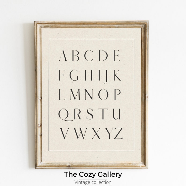 May include: A framed print of the alphabet in a simple, modern font. The print is on a beige background and has a light wood frame. The text at the bottom of the print reads "The Cozy Gallery, Vintage Collection."