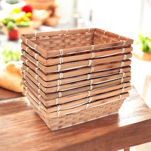 May include: A stack of rectangular woven bamboo baskets. The baskets are a light brown colour and are held together with white string. The baskets are sitting on a wooden surface. The background is blurred, but appears to be a kitchen.