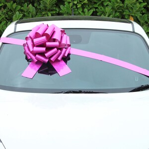 May include: A white car with a large pink bow on the windscreen. The bow is made of shiny, metallic ribbon.