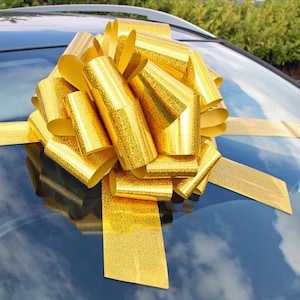 May include: A large gold bow with a glittery finish. The bow is tied on top of a car.