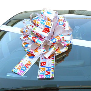 May include: A white car bow with a colourful "Happy Birthday" design. The bow is made of white ribbon with colourful printed text and stars. The bow is tied on the front of a car.