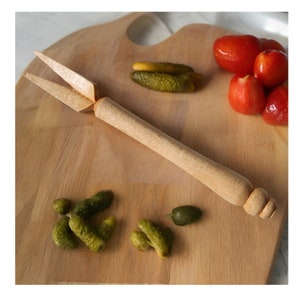 May include: A wooden pickle fork with a long handle on a wooden chopping board with green pickles and red tomatoes.