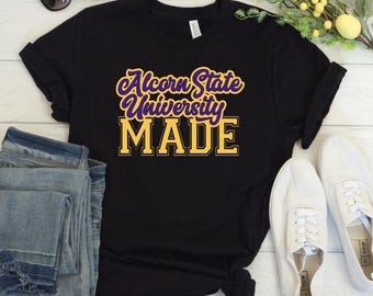 May include: Black t-shirt with the text "Alcorn State University MADE" in purple and gold lettering. The shirt is folded, with blue jeans and white sneakers visible in the background.