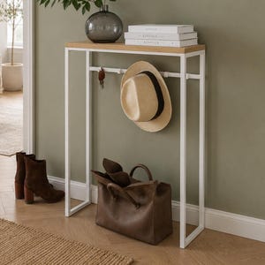 Può includere: Un tavolo consolle in metallo bianco con piano in legno chiaro, dotato di una barra orizzontale con ganci per appendere oggetti. Un cappello di paglia è appeso a un gancio, mentre una borsa in pelle marrone è appoggiata sotto. Un vaso in vetro.