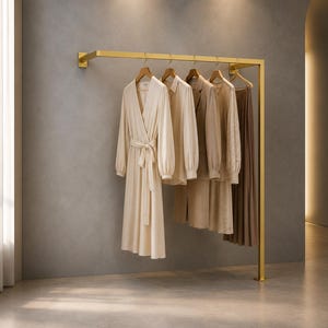 Pode incluir: Um cabideiro de parede em metal dourado com uma barra horizontal suportada por um poste vertical. Quatro cabides de madeira sustentam um vestido envelope na cor creme, uma camisa bege claro, um blazer bege e uma saia plissada.