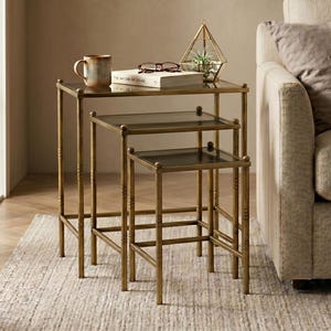 Op de afbeelding: Een set van drie nesttafels met een goudkleurig metalen frame en glazen bladen. De tafels zijn trapsgewijs gerangschikt, met de grootste tafel bovenaan en de kleinste onderaan. Een mok, boek en terrarium staan op de bovenste tafel.