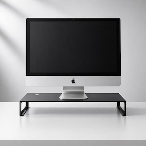 May include: A silver iMac computer with a black screen sits on a black monitor stand. The stand is rectangular with a small hole on the top surface. The Apple logo is visible on the computer.