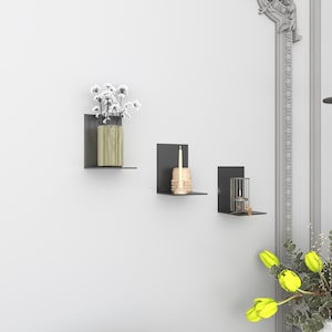 May include: Three black metal floating shelves mounted on a white wall. The top shelf holds a vase with white flowers. The middle shelf holds a candle and the bottom shelf holds a candle holder.
