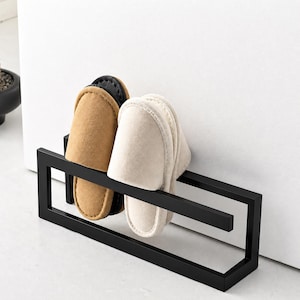 Black metal wall mounted slipper holder rack with minimalist geometric design. Matte finish shoe organizer storing two pairs of indoor slippers. Modern space saving wall storage solution for entryway, bathroom, or home organization.