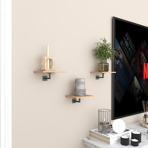 May include: Three wooden floating shelves with black metal brackets are mounted on a beige wall. Each shelf holds a decorative item: a candle, a vase, and a small plant in a glass vase. The shelves are a simple, modern design.