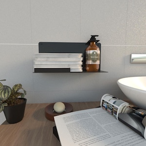 Puede incluir: Una estantería de metal negro montada en una pared de azulejos grises, que contiene una pila de revistas blancas y una botella marrón con una bomba negra. Una pequeña planta en maceta y un jabonera de madera con jabón blanco están sobre una superficie de madera.