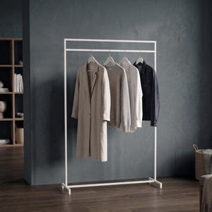 May include: A white metal clothing rack with a rectangular frame, holding four garments on hangers: a long beige coat, a grey sweater, a white button-down shirt, and a dark navy blue jacket, all displayed in a row on the horizontal bar.