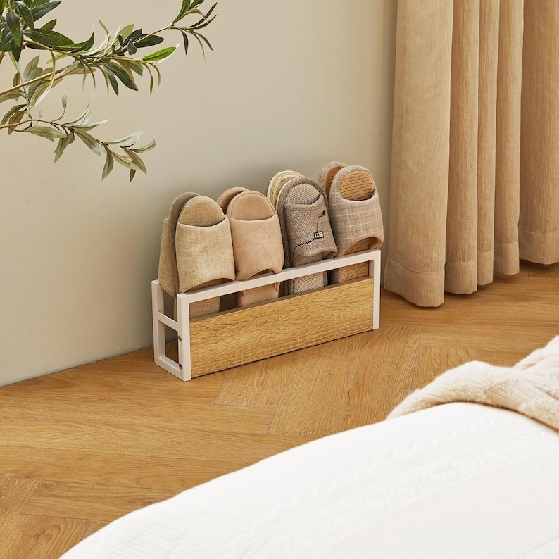 Puede incluir: Un zapatero de madera y metal blanco que contiene varios pares de zapatillas. Las zapatillas son de varios colores neutros, incluyendo beige, beige claro y marr&oacute;n. El zapatero est&aacute; colocado sobre un suelo de madera, con una pared y una cortina beige en el fondo.
