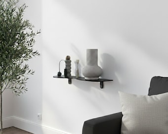 May include: A black, wavy-edged wall shelf displays decorative items. A gray vase, a small sculpture, and other objects are arranged on the shelf. The shelf is mounted on a white wall, with a tree and a couch visible.