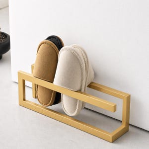 Matte gold metal wall mounted slipper holder with minimalist geometric design. Modern shoe organizer rack holding two pairs of indoor slippers. Space saving wall storage solution for entryway, bathroom, or home organization.