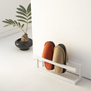 May include: A white metal shoe rack with two pairs of slippers. One pair is orange and the other is beige.