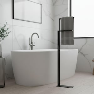 Pode incluir: Um toalheiro de metal preto com uma toalha cinza pendurada. O toalheiro está em um piso de madeira clara em um banheiro. Uma banheira branca e uma janela estão ao fundo.