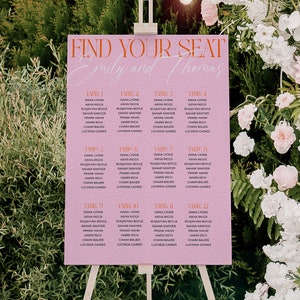May include: Pink wedding seating chart with the text "Find Your Seat" and "Emily and Thomas" in orange. The chart is on an easel in front of a floral arrangement.