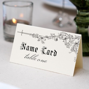 Może przedstawiać: Kremowa wizytówka z czarnymi ozdobami i napisami "Name Card table one". Na górze znajduje się wzór miecza. Karta leży na białym obrusie, z zapaloną świecą i zielonym szkłem w tle.