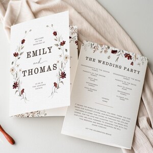 May include: Wedding stationery set featuring a floral design with burgundy and white flowers. The invitation reads "Emily and Thomas" with the wedding date and venue. Includes a wedding party program and wax seals.
