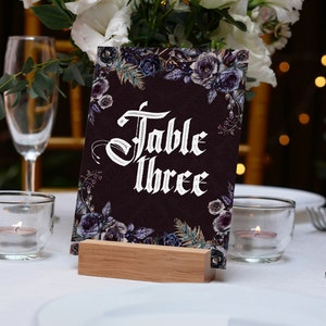 Op de afbeelding: Een tafelnummerkaart met de tekst "Table Three" in witte gotische letters, omringd door bloemmotieven in donkerpaars en blauw. De kaart staat op een houten blok. De sc&egrave;ne omvat kaarsen, glazen en een wit tafelkleed, geschikt voor een bruiloft of evenement.