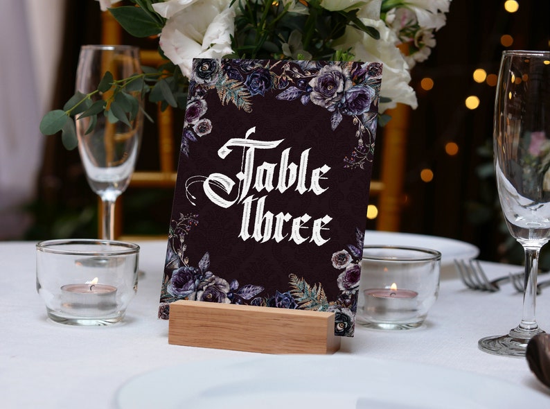 Op de afbeelding: Een tafelnummerkaart met de tekst "Table Three" in witte gotische letters, omringd door bloemmotieven in donkerpaars en blauw. De kaart staat op een houten blok. De sc&egrave;ne omvat kaarsen, glazen en een wit tafelkleed, geschikt voor een bruiloft of evenement.
