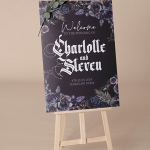May include: A black welcome sign with white gothic lettering announcing the wedding of Charlotte and Steven. The sign is decorated with dark purple and blue floral illustrations and the date "4th July 2024" and the location "Harelaw Farm". The sign is displayed on a wooden easel.