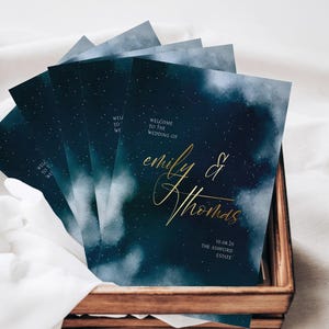 May include: Wedding welcome cards with a dark blue watercolour design and gold script. The cards read "Welcome to the Wedding of Emily & Thomas" and include the date and venue.