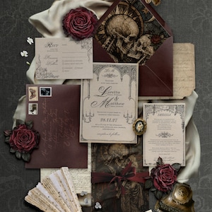 May include: A wedding stationery set featuring cream-colored cards with black gothic-style text, dark burgundy envelopes with skull illustrations, deep red roses, a lace folding fan, and a vintage-style brass inkwell on a dark gray surface.
