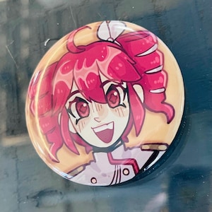 May include: A circular badge featuring a cartoon character with pink hair and eyes, smiling widely. The badge has a glossy finish and a light yellow background. The character is wearing a white uniform.