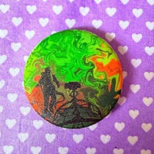 May include: A circular pinback button featuring a black silhouette of a person standing near a tripod-mounted camera against a swirling, glittery background of vibrant neon green, orange, and dark red colors that resemble a nebula or gas cloud.
