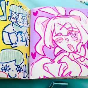 May include: A sketchbook open to two colorful pages of cartoon illustrations. One page features a yellow background with a blue and green drawing of a person. The other page has a purple background with a pink drawing of a girl with hearts.