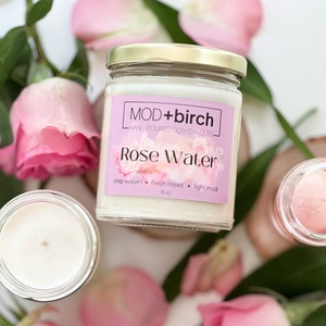 May include: A glass jar candle with a gold lid. The label reads "MOD+birch HAND POURED SOY CANDLES Rose Water crisp waters + fresh roses + light musk 9 oz."