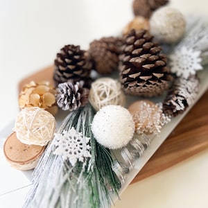 May include: A white tray with a winter-themed arrangement of pine cones, white pinecones, white snowflakes, and white and brown balls. The tray is on a wooden surface.