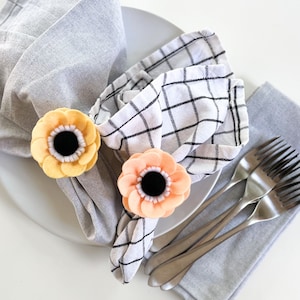 May include: Two felt flower napkin rings, one yellow and one peach, are placed on white and gray checkered napkins. The napkins are folded and placed on a white plate.