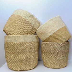 Puede incluir: Cuatro cestas tejidas de color beige natural con una franja blanca alrededor del centro. Las cestas están apiladas una encima de la otra.