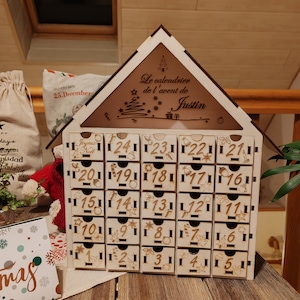May include: Wooden advent calendar in the shape of a house. It has 24 small drawers, each numbered and decorated with festive symbols. The top section has text that reads "Le calendrier de l'avent de Justin".