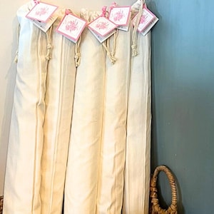 May include: Several cream-colored fabric items are displayed, each with a tag attached by a pink ribbon. The tags have a floral design. The items are neatly arranged, and a woven basket is visible in the foreground.