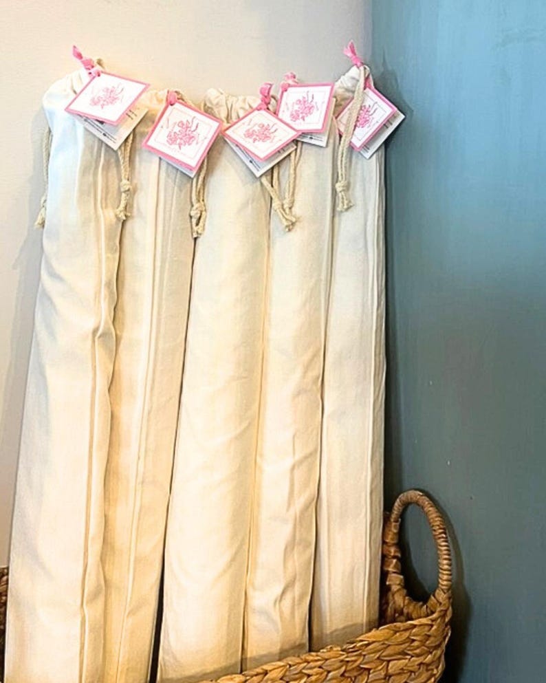 May include: Several cream-colored fabric items are displayed, each with a tag attached by a pink ribbon. The tags have a floral design. The items are neatly arranged, and a woven basket is visible in the foreground.