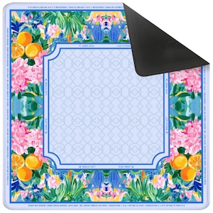 May include: A colorful, rectangular play mat with a floral and fruit design. The mat features oranges, pink flowers, and a light blue background with a geometric pattern. The edges have text and a blue border. The back of the mat is black.