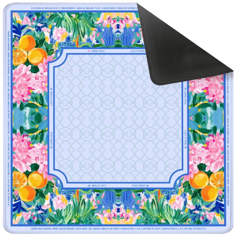 May include: A colorful, rectangular play mat with a floral and fruit design. The mat features oranges, pink flowers, and a light blue background with a geometric pattern. The edges have text and a blue border. The back of the mat is black.