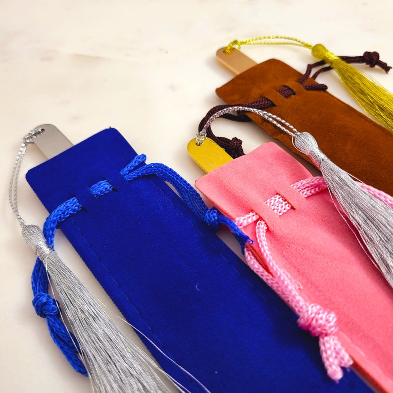 May include: Four velvet bookmark pouches with tassels. The pouches are blue, pink, brown, and gold. The tassels are silver, white, and yellow.