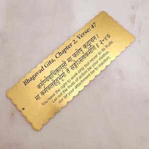May include: A gold bookmark with a Sanskrit quote from the Bhagavad Gita, Chapter 2, Verse 47. The quote reads: "Karmanyevaadhikaarastee maa phaleshu kadaachana. Maa karmaphalahetoorbhoormaa te sangostvakarmani." The English translation is: "You have the right to work only but never to its fruits. Let not the fruits of action be your motive, nor let your attachment be to inaction."