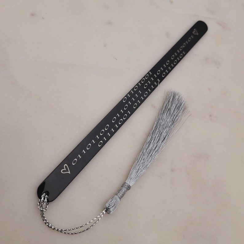 May include: A black bookmark with a silver tassel. The bookmark is engraved with a heart and a series of 0s and 1s.
