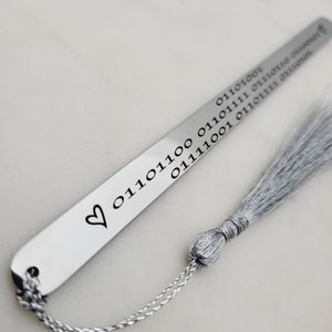 May include: A silver bookmark with a heart and a string of binary code engraved on it. The bookmark has a silver tassel.