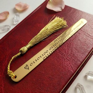 May include: A gold-colored metal bookmark with a binary code pattern and a heart symbol. The bookmark has a gold tassel attached. The bookmark is resting on a red leather-bound book.