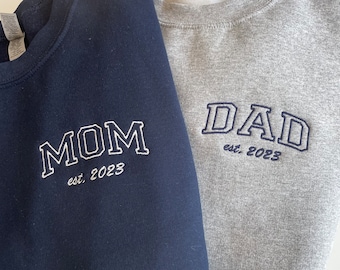 May include: Two navy blue and gray sweatshirts with embroidered text. The navy blue sweatshirt says "MOM est. 2023" and the gray sweatshirt says "DAD est. 2023".