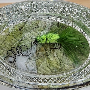 May include: A clear glass oval dish with a textured rim. The dish contains a green and yellow fly fishing lure with black thread.